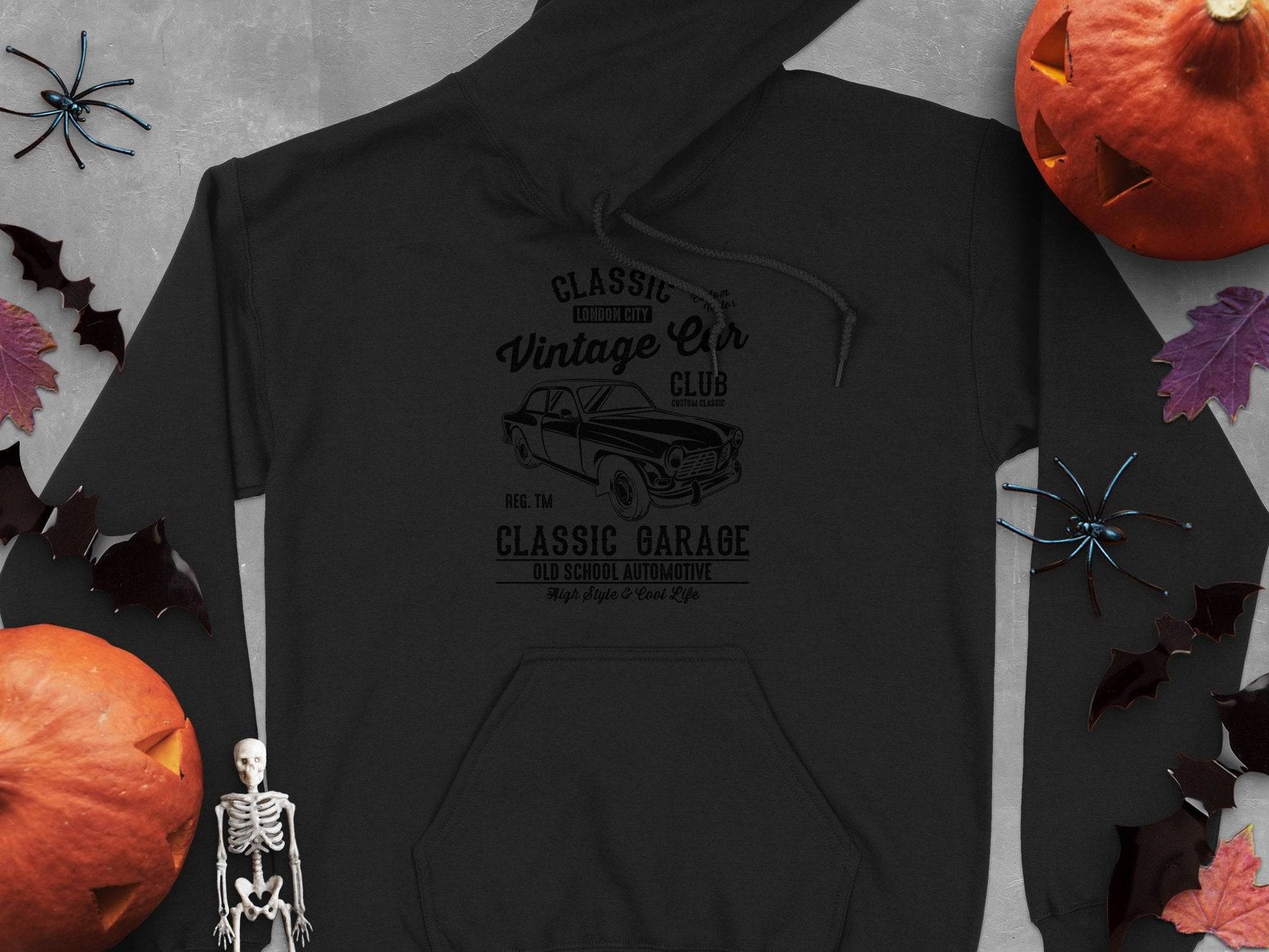 The black hoodie by Garment Graphics has a classic fit with a vintage car graphic and Classic Car Club and Classic Garage text, surrounded by Halloween elements like pumpkins, bats, leaves, and a skeleton—ideal for car enthusiasts.