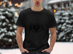 In a snowy outdoor setting, a person wears a black Garment Graphics T-shirt with Scanir 8 Power and intricate designs. Snow-covered trees blur in the background as they casually tuck their hands into pockets.