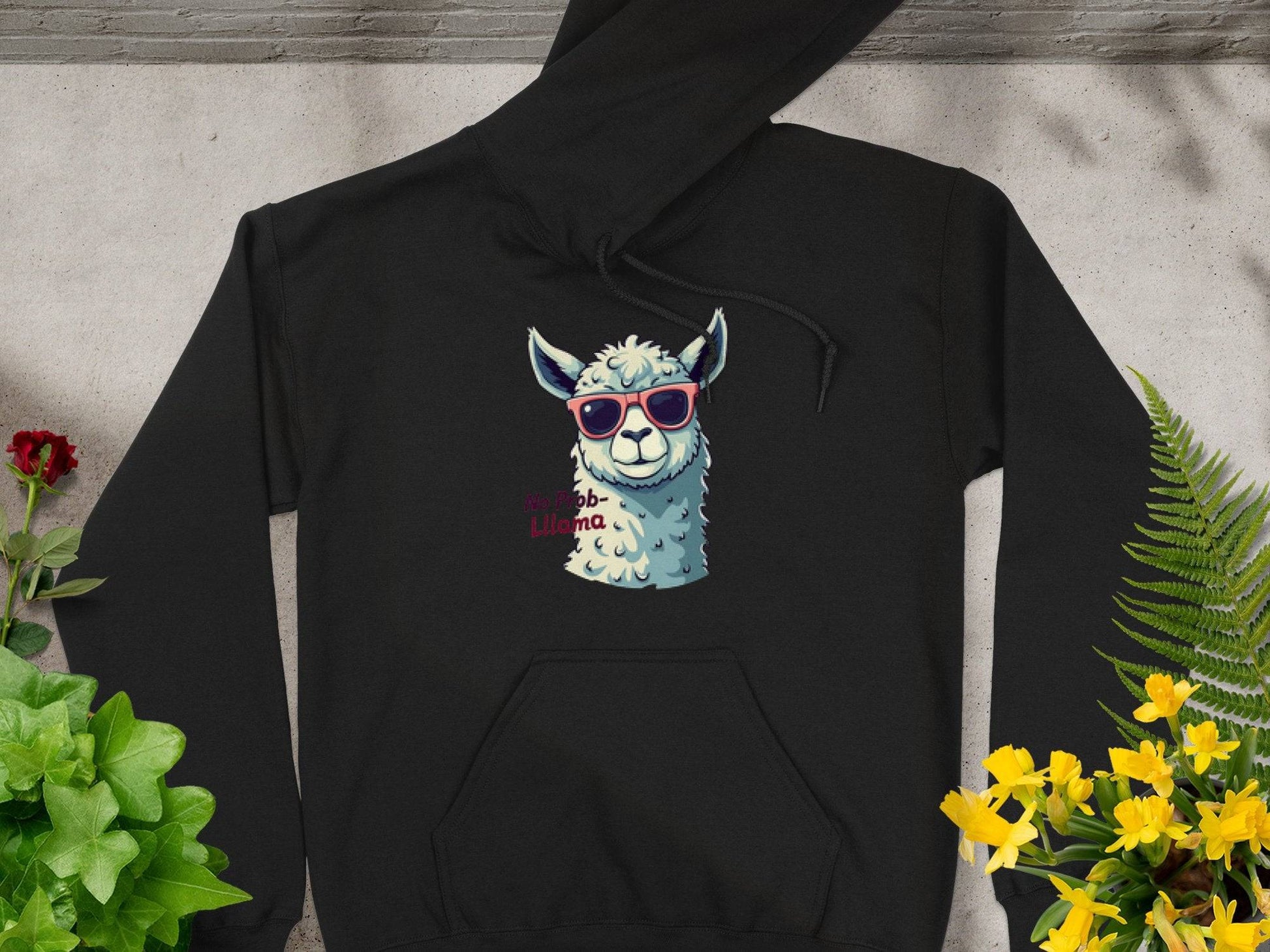 A classic fit black hoodie from Garment Graphics is displayed on a gray surface. It features a llama in funny red sunglasses and probllama in pink text, surrounded by yellow flowers and green plants.