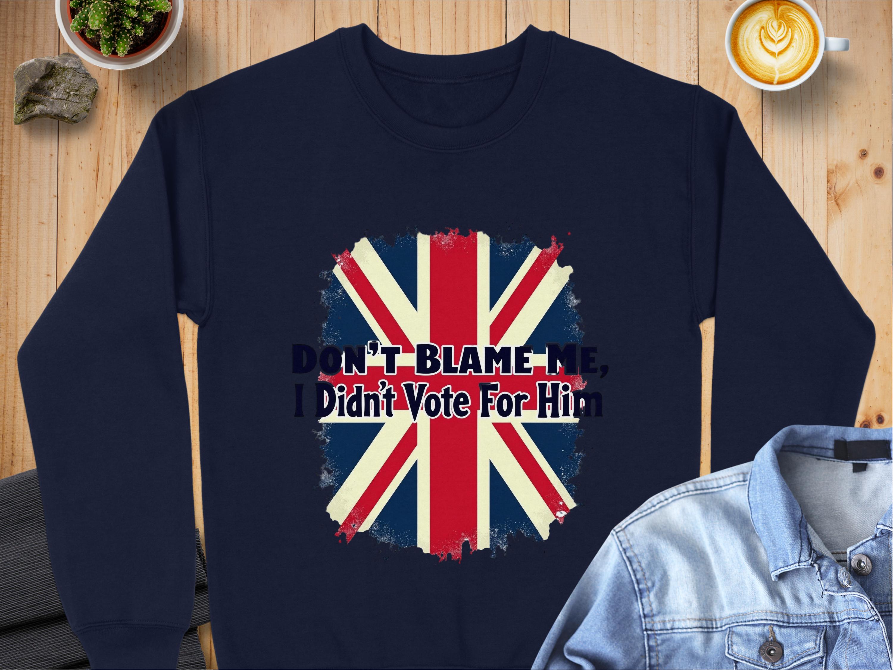 A navy unisex sweatshirt by Garment Graphics features a distressed Union Jack and the phrase DONT BLAME ME, I Didnt Vote For Him, styled on a wooden table with a denim jacket and steaming latte.