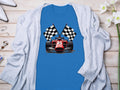A Garment Graphics blue T-shirt with a red Formula 1 car and two checkered flags, laid on wood, paired with a white cardigan, chic heels, and small white flowers—a perfect motorsport enthusiasts ensemble.