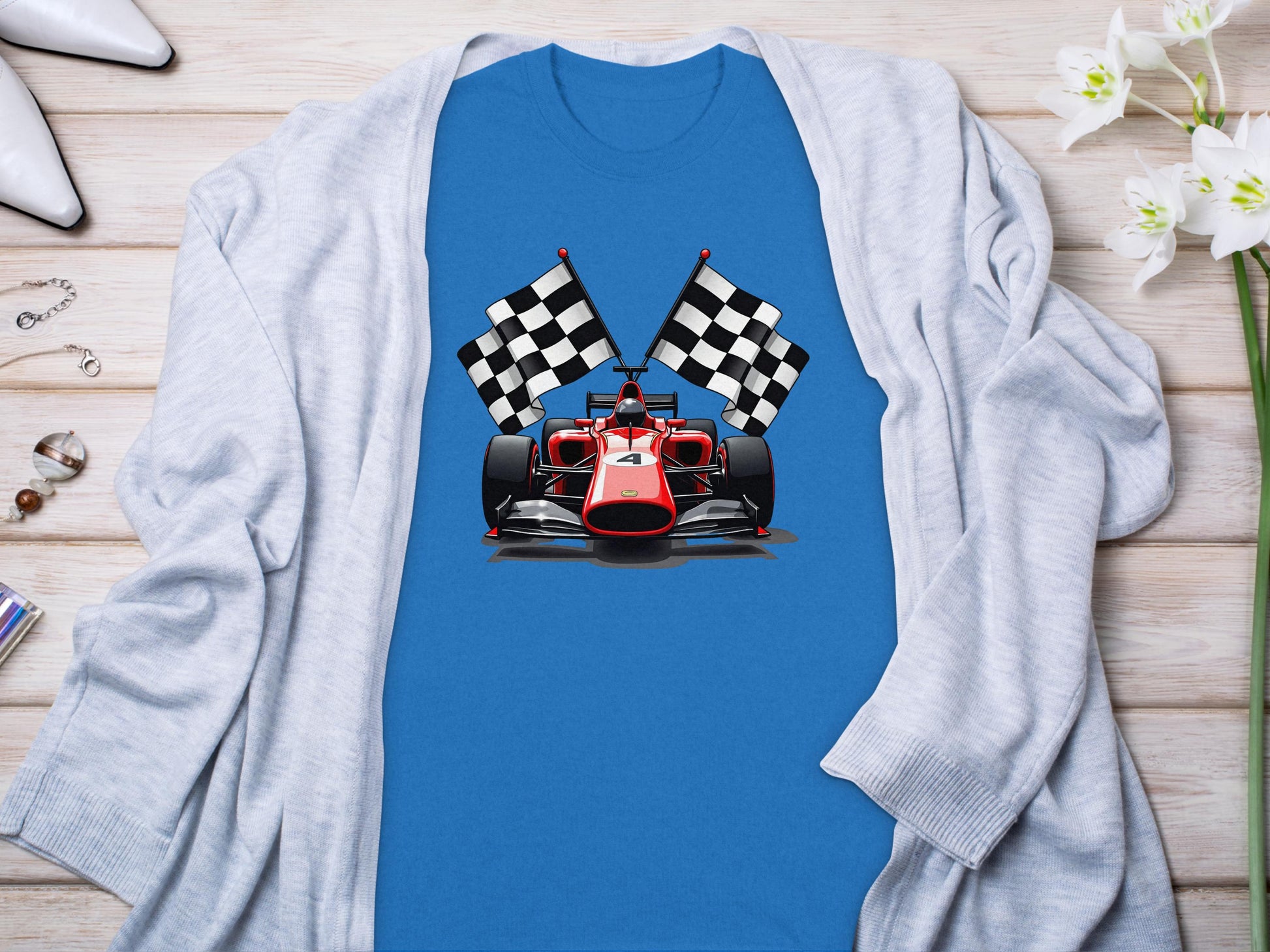 A Garment Graphics blue T-shirt with a red Formula 1 car and two checkered flags, laid on wood, paired with a white cardigan, chic heels, and small white flowers—a perfect motorsport enthusiasts ensemble.