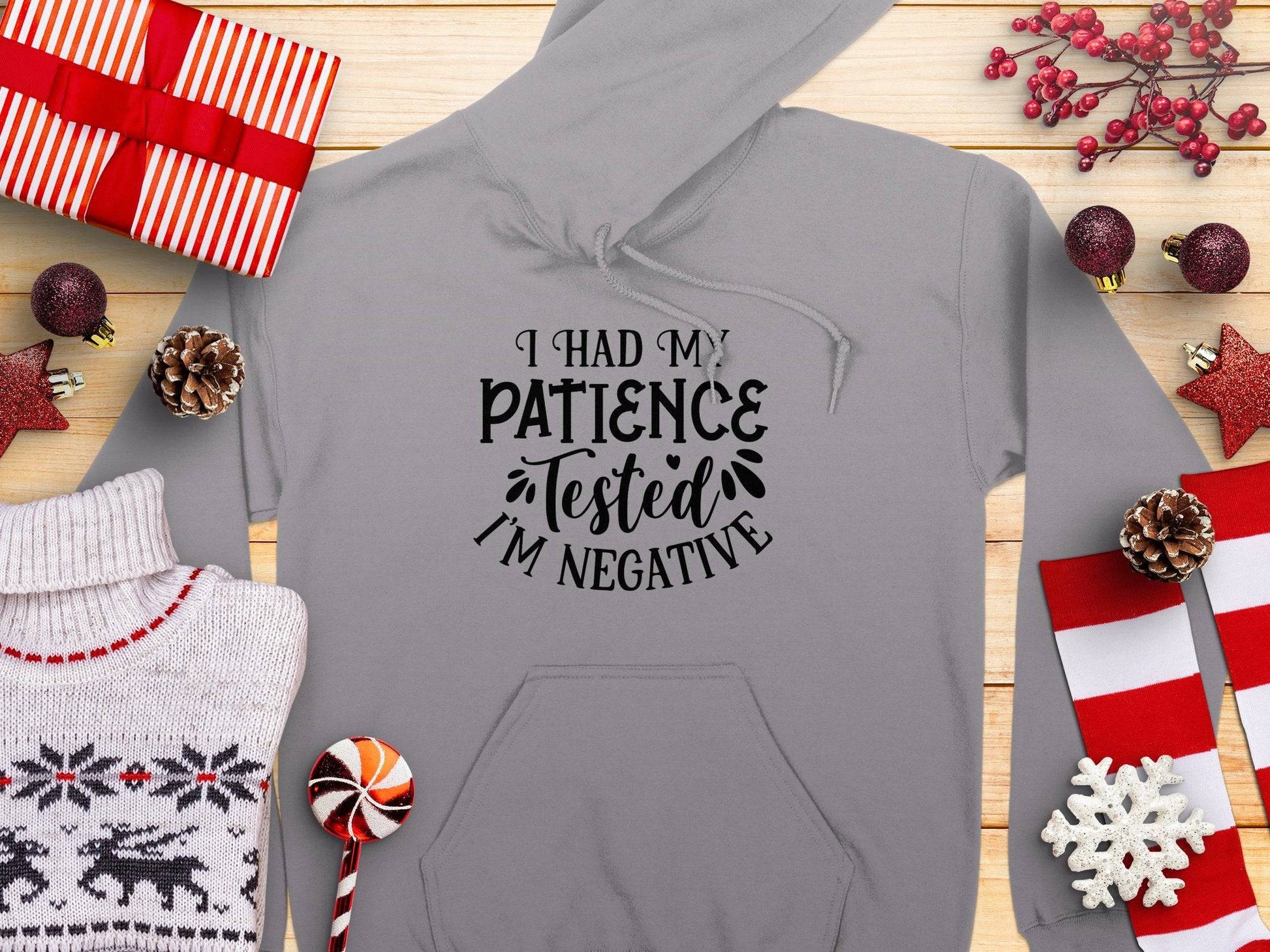 A Garment Graphics Patience Tested Hoodie, featuring the text I had my patience tested Im negative in gray, is surrounded by gifts, a classic fit sweater, pine cones, candy, and Christmas decorations on wood. This medium-heavy fabric hoodie is ideal for the festive season.