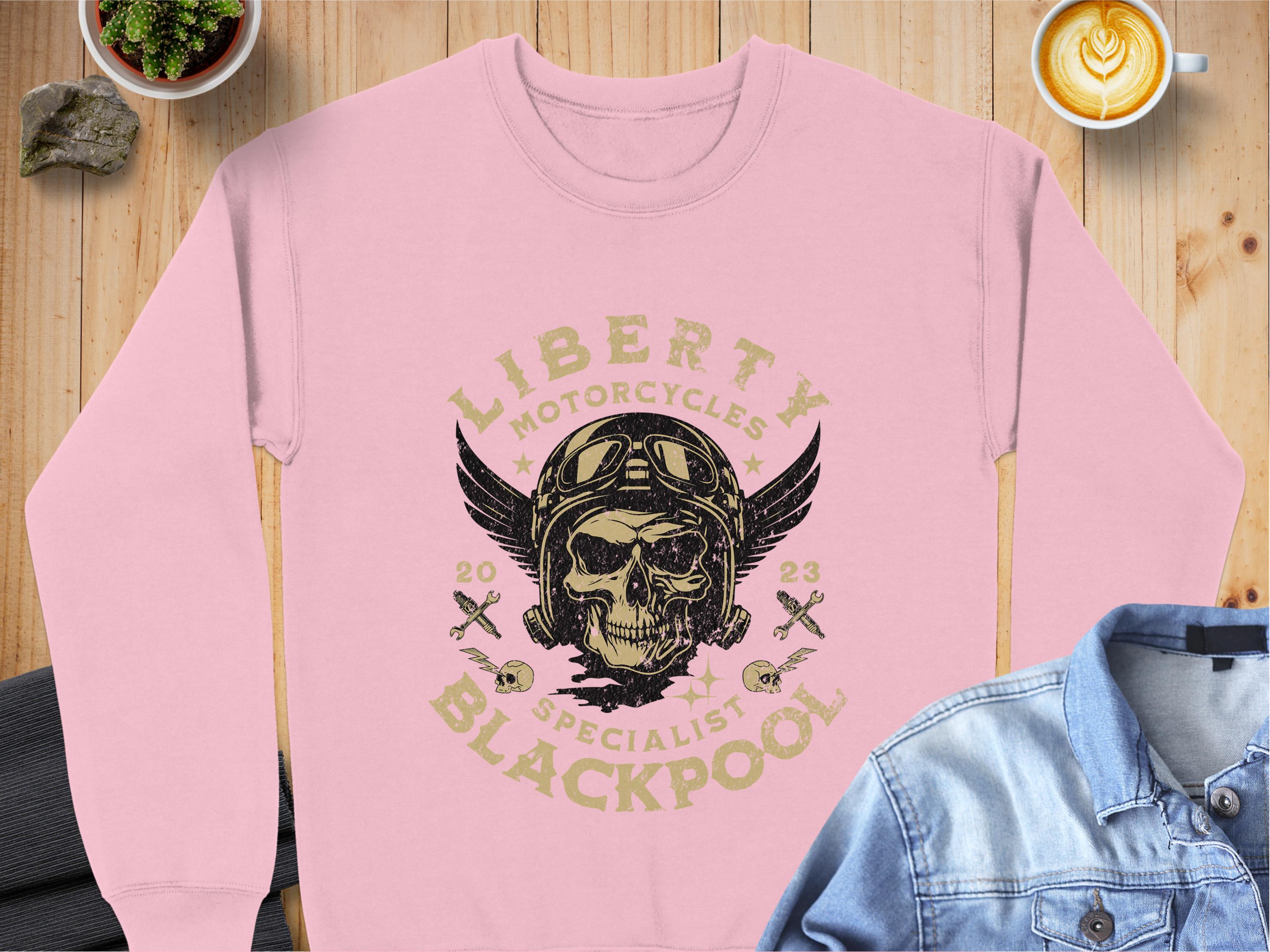 The Garment Graphics pink Blackpool 2023 Sweatshirt features a skull in goggles with a winged helmet and the text Liberty Motorcycles Specialist Blackpool 2023. It is displayed on a wooden table, paired with a denim shirt and accompanied by a latte.