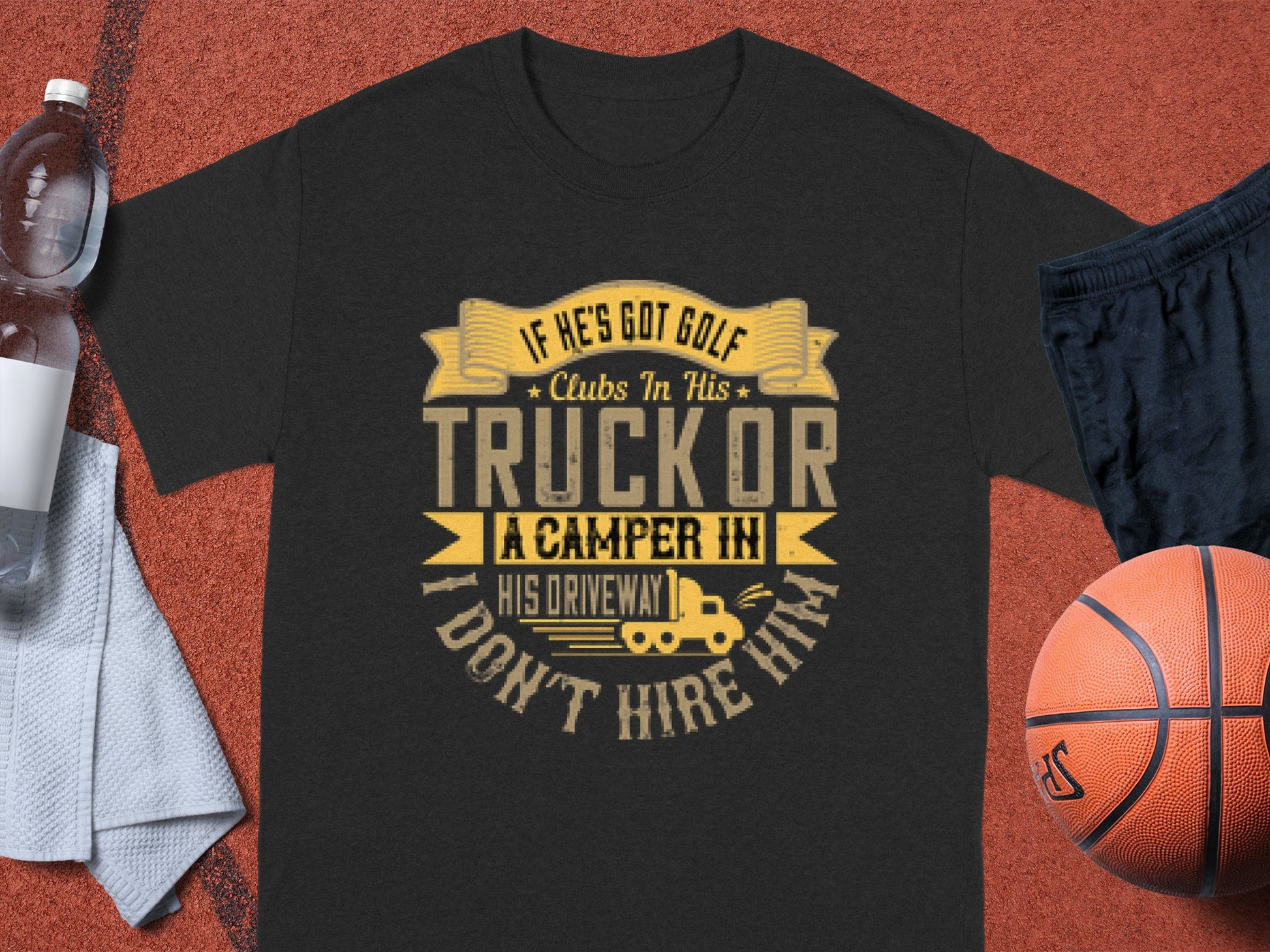 A black Garment Graphics t-shirt lies on a reddish-brown surface with yellow and beige text: If hes got golf clubs in his truck or a camper in his driveway, I dont hire him. Nearby are a water bottle, light gray towel, and orange basketball, adding a touch of golf and camping vibe.