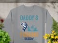 Garment Graphics presents the ideal gift for fishing fans: a gray sweatshirt featuring Daddys Fishing Buddy in vibrant teal and orange. It showcases a fish and person fishing on a boat amid green and yellow foliage on wood.