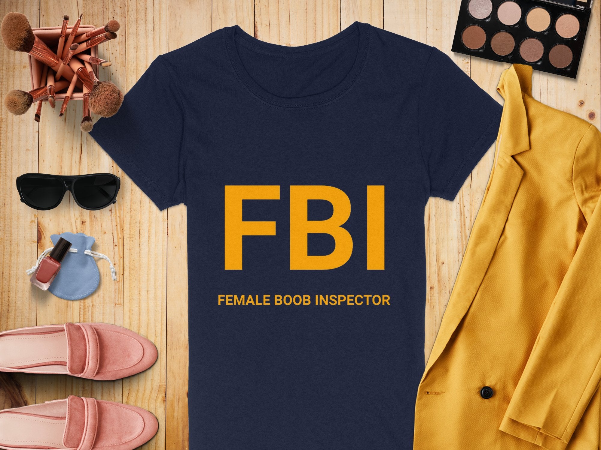 A navy Garment Graphics FBI FEMALE BOOB INSPECTOR T-shirt, perfect as a gag gift and available in womens sizing, is on a wooden surface surrounded by makeup brushes, an eyeshadow palette, sunglasses, nail polish, pink shoes, a coin purse, and a yellow blazer.