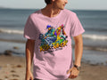A person sports a Garment Graphics pink t-shirt showcasing an artistic design of two dancers, one blue and the other pink, alongside the phrases Risk Taker and Street Dance with Flava, creating a dynamic nod to street dance culture against the ocean backdrop.