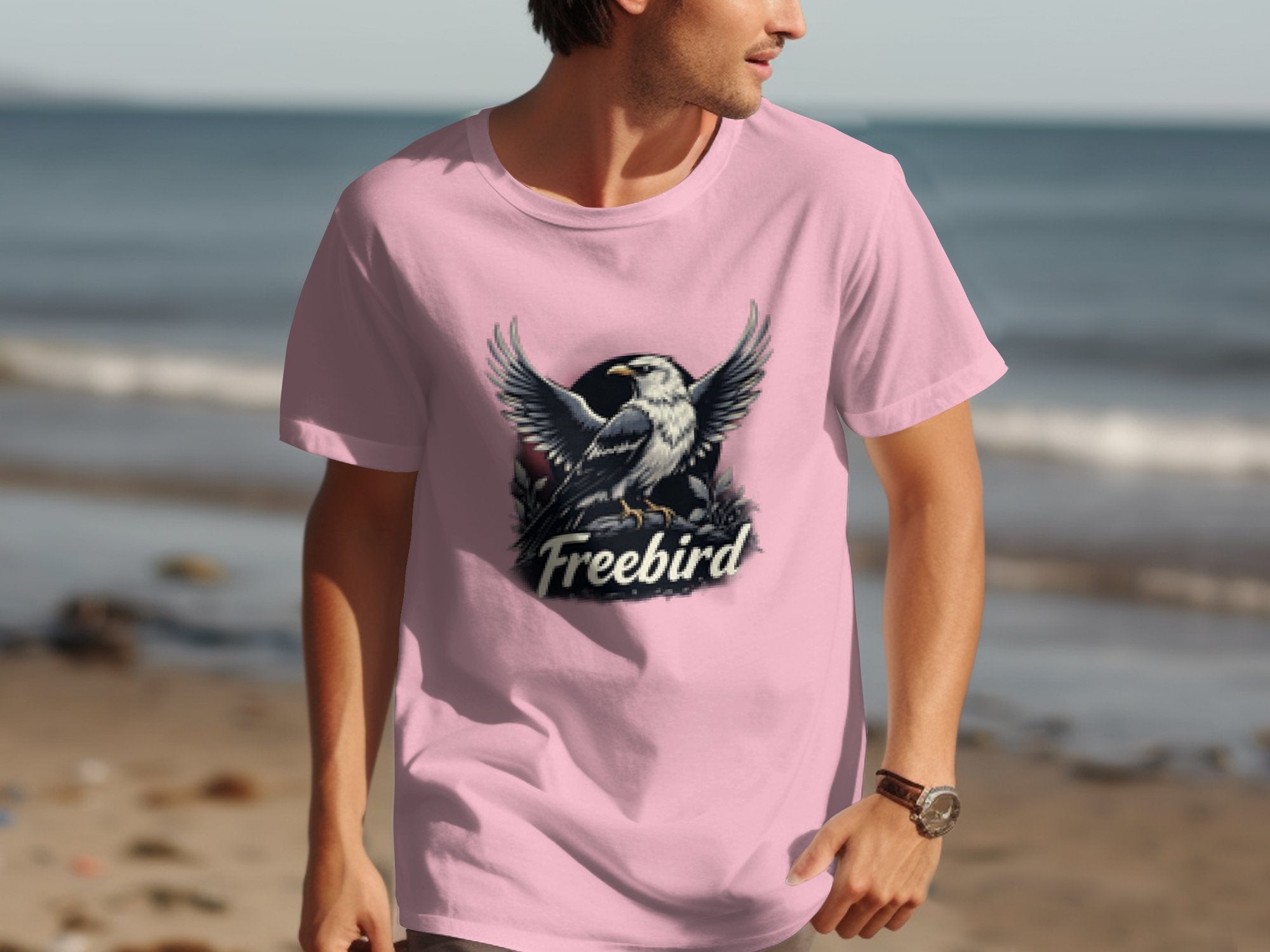 A person strolls along the beach in a light pink Garment Graphics shirt, made of 100% cotton. The bold eagle graphic stands out against the sunny backdrop as sand and azure water complete this idyllic scene under a clear sky.