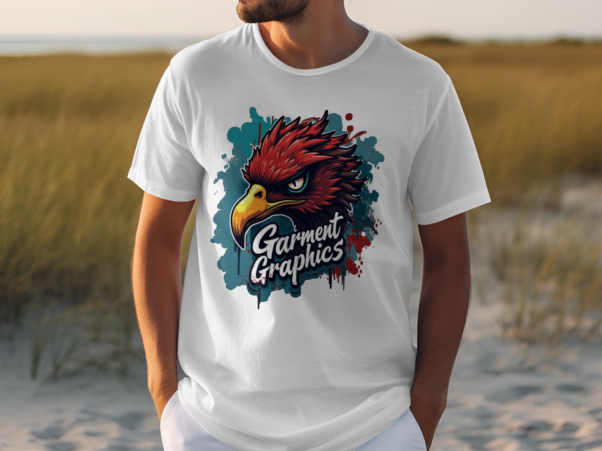 A person in a white Garment Graphics T-shirt with a colorful eagle design stands outdoors amidst grassy dunes.