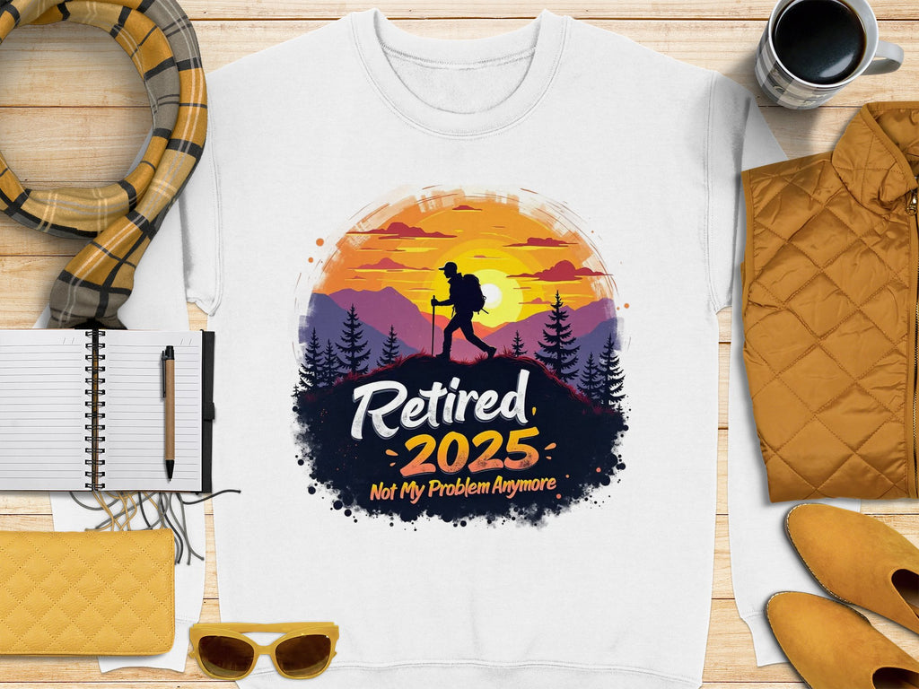 Garment Graphics offers a white unisex T-shirt with a hiker design showing tree and mountain silhouettes against a sunset, featuring the text Retired 2025 Not My Problem Anymore. Pair it with items like a scarf, notebook, pen, coffee cup, quilted bag, sunglasses, or yellow jacket.