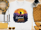 Garment Graphics offers a white unisex T-shirt with a hiker design showing tree and mountain silhouettes against a sunset, featuring the text Retired 2025 Not My Problem Anymore. Pair it with items like a scarf, notebook, pen, coffee cup, quilted bag, sunglasses, or yellow jacket.