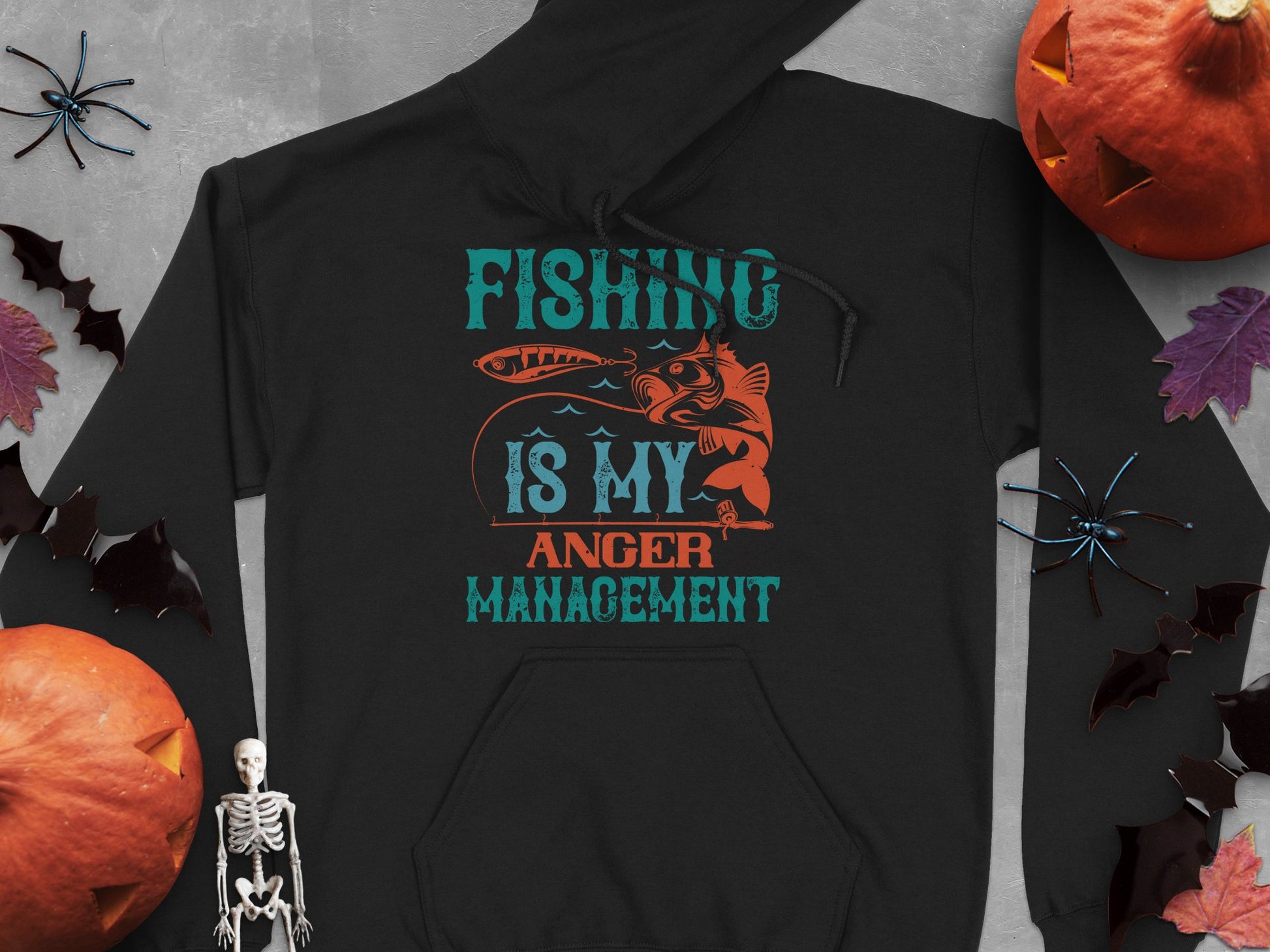 Garment Graphics offers a black hoodie with Fishing Is My Anger Management text and a fish illustration, surrounded by Halloween-themed items like pumpkins, bats, and a small skeleton on a textured gray surface.