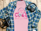 Garment Graphics pink shirt with Just be Cool Rock and Roll in purple and gray, perfect for music lovers, with headphones. It rests on a plaid blue-green flannel. Surrounding items: a camera, headphones, and a smartphone on a wooden surface.