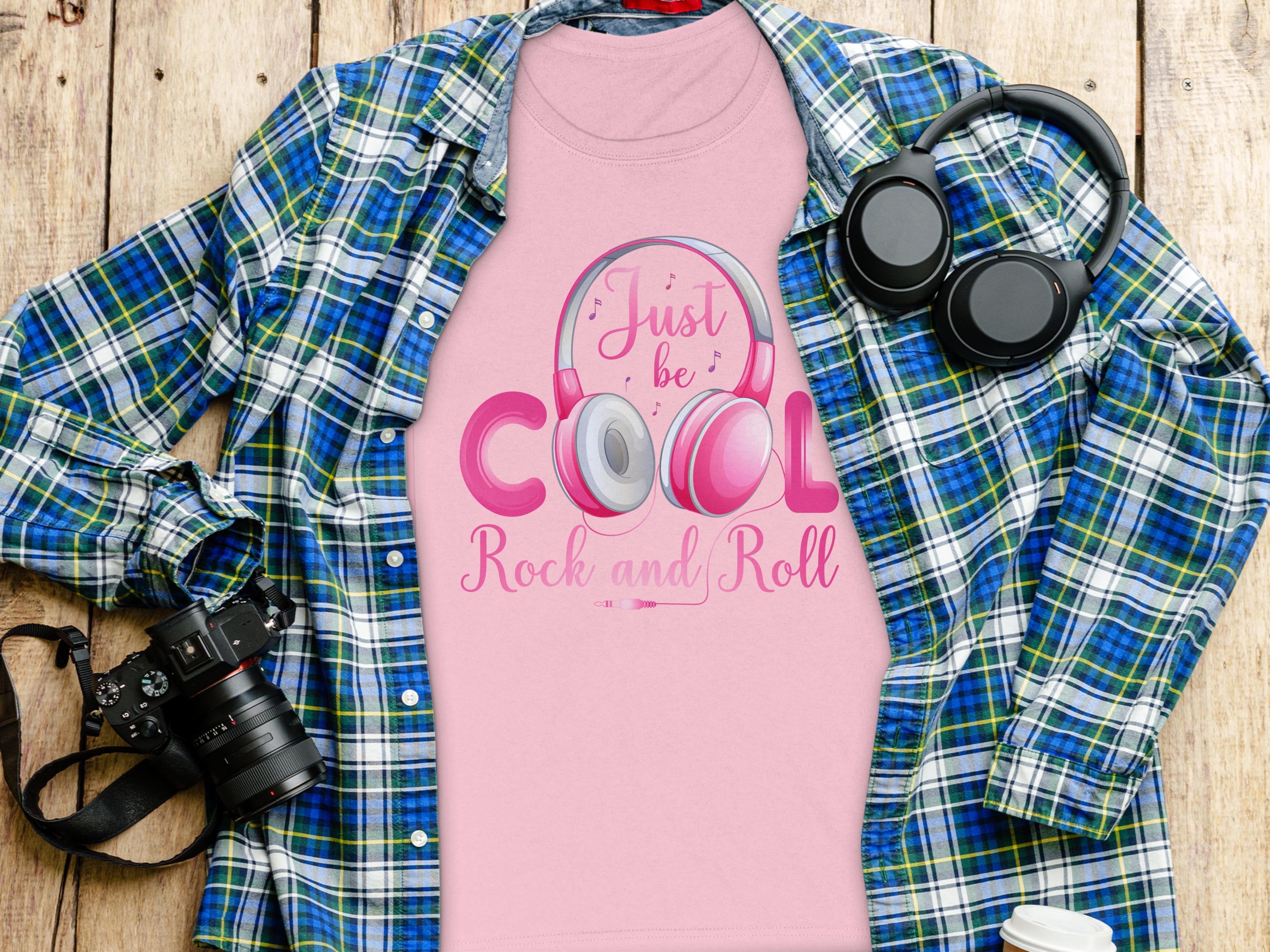 Garment Graphics pink shirt with Just be Cool Rock and Roll in purple and gray, perfect for music lovers, with headphones. It rests on a plaid blue-green flannel. Surrounding items: a camera, headphones, and a smartphone on a wooden surface.