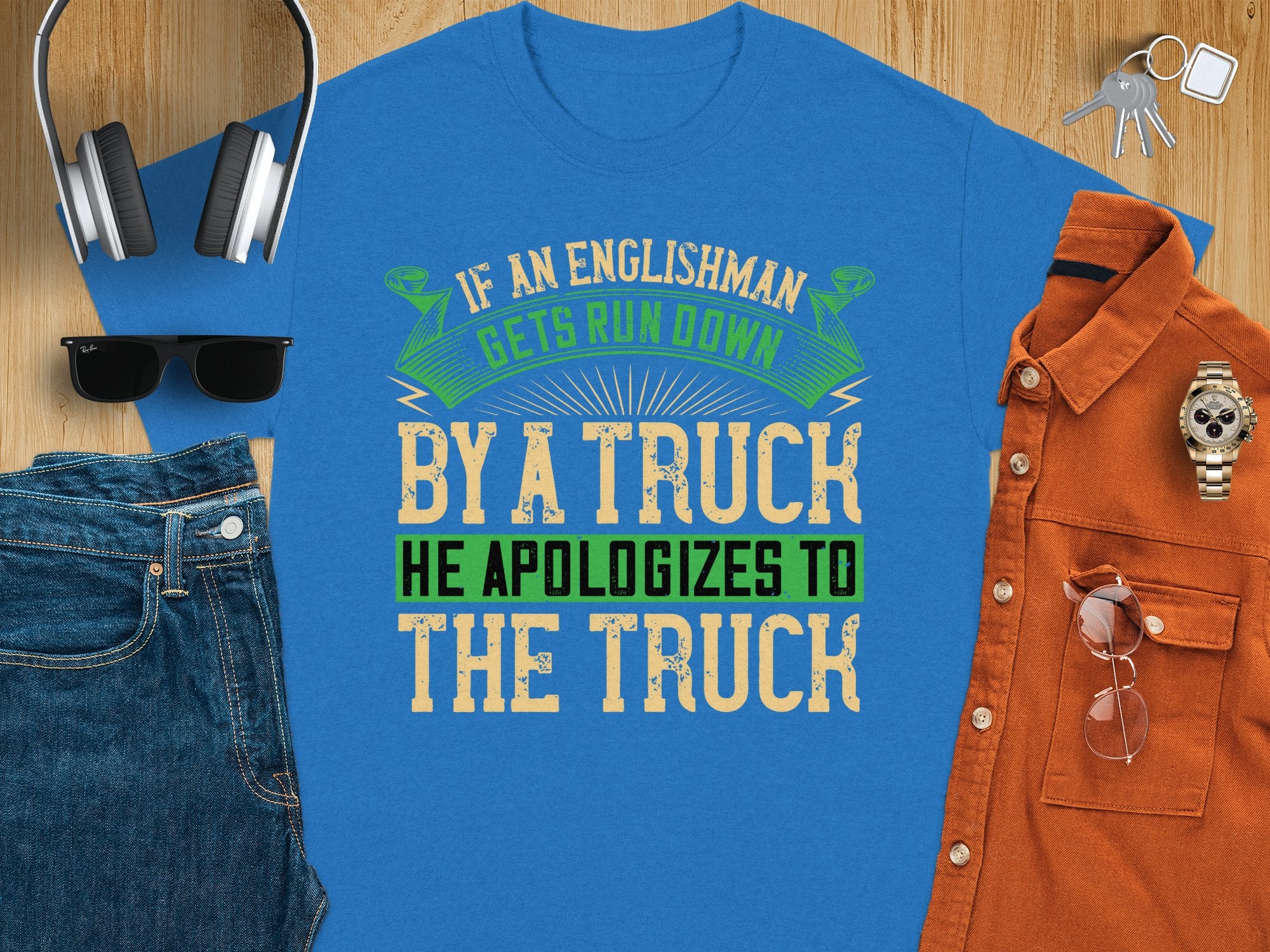 A Garment Graphics blue t-shirt, featuring the witty text If an Englishman gets run down by a truck he apologizes to the truck, is displayed on wood with a brown shirt, jeans, sunglasses, a watch, headphones, and a blue wallet.