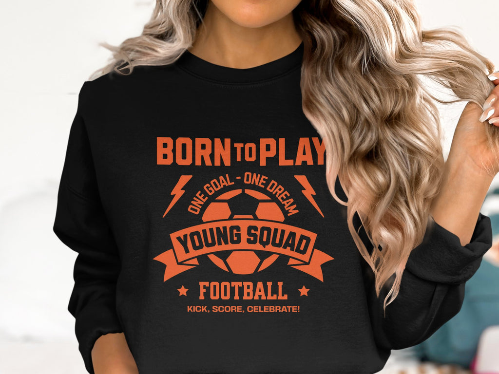 A person wearing a Garment Graphics unisex black football sweatshirt showcases an orange soccer ball graphic with the bold text BORN TO PLAY, ONE GOAL - ONE DREAM, YOUNG SQUAD FOOTBALL, KICK, SCORE, CELEBRATE! Their long, wavy hair obscures their face, revealing only their torso.