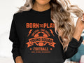 A person wearing a Garment Graphics unisex black football sweatshirt showcases an orange soccer ball graphic with the bold text BORN TO PLAY, ONE GOAL - ONE DREAM, YOUNG SQUAD FOOTBALL, KICK, SCORE, CELEBRATE! Their long, wavy hair obscures their face, revealing only their torso.