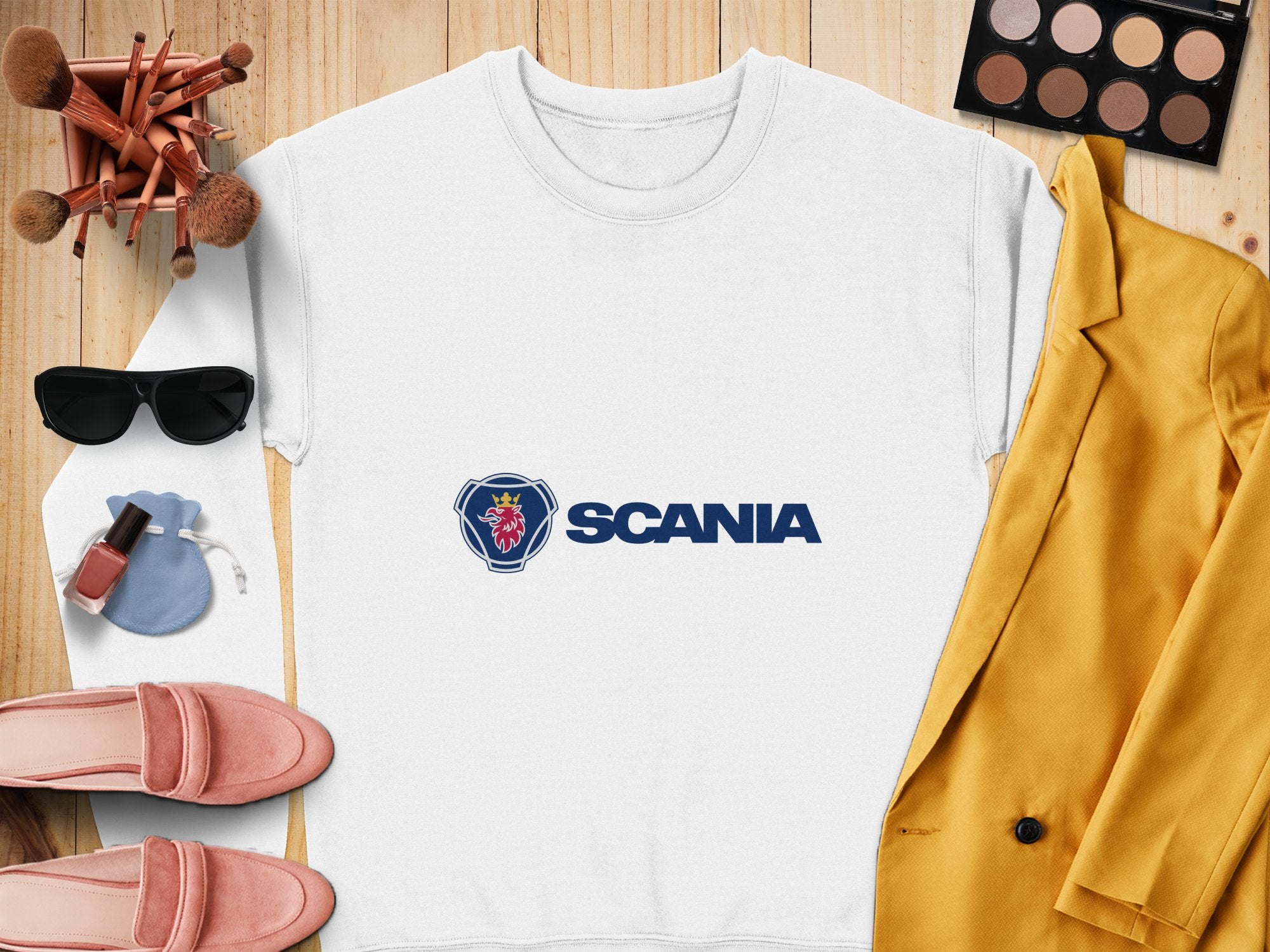 A white Scania logo sweatshirt by Garment Graphics, ideal for casual outings, is displayed on a wooden surface alongside a yellow jacket, pink shoes, sunglasses, makeup brushes, an eyeshadow palette, nail polish, and a small blue pouch. Unisex sizing offers effortless style daily.