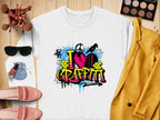 A white t-shirt from Garment Graphics featuring a vibrant graffiti design is displayed on a wooden surface, radiating urban style. Around it are sunglasses, makeup brushes, a palette, lipstick, pink shoes, a light blue pouch, and a yellow blazer.