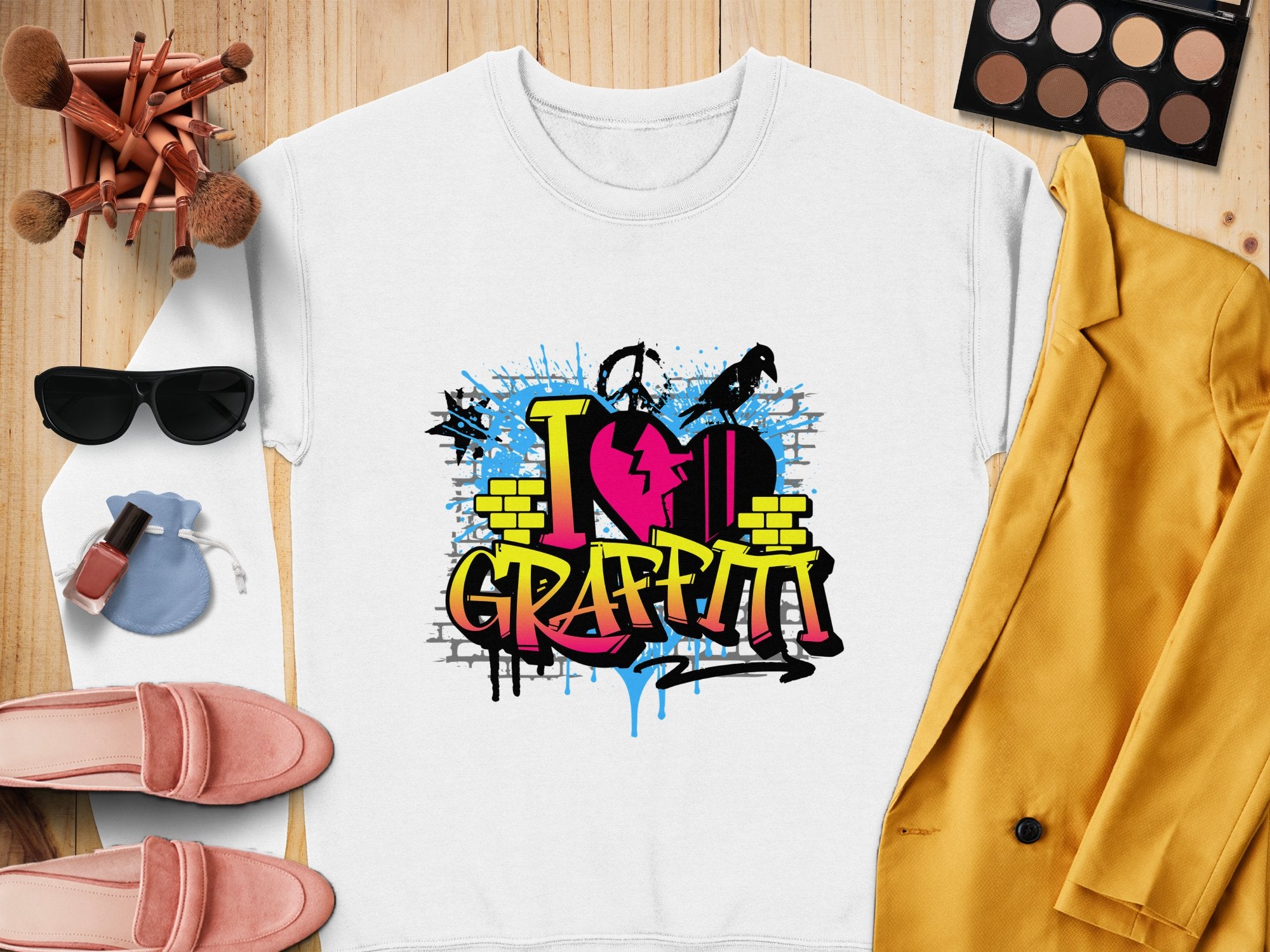 A white t-shirt from Garment Graphics featuring a vibrant graffiti design is displayed on a wooden surface, radiating urban style. Around it are sunglasses, makeup brushes, a palette, lipstick, pink shoes, a light blue pouch, and a yellow blazer.