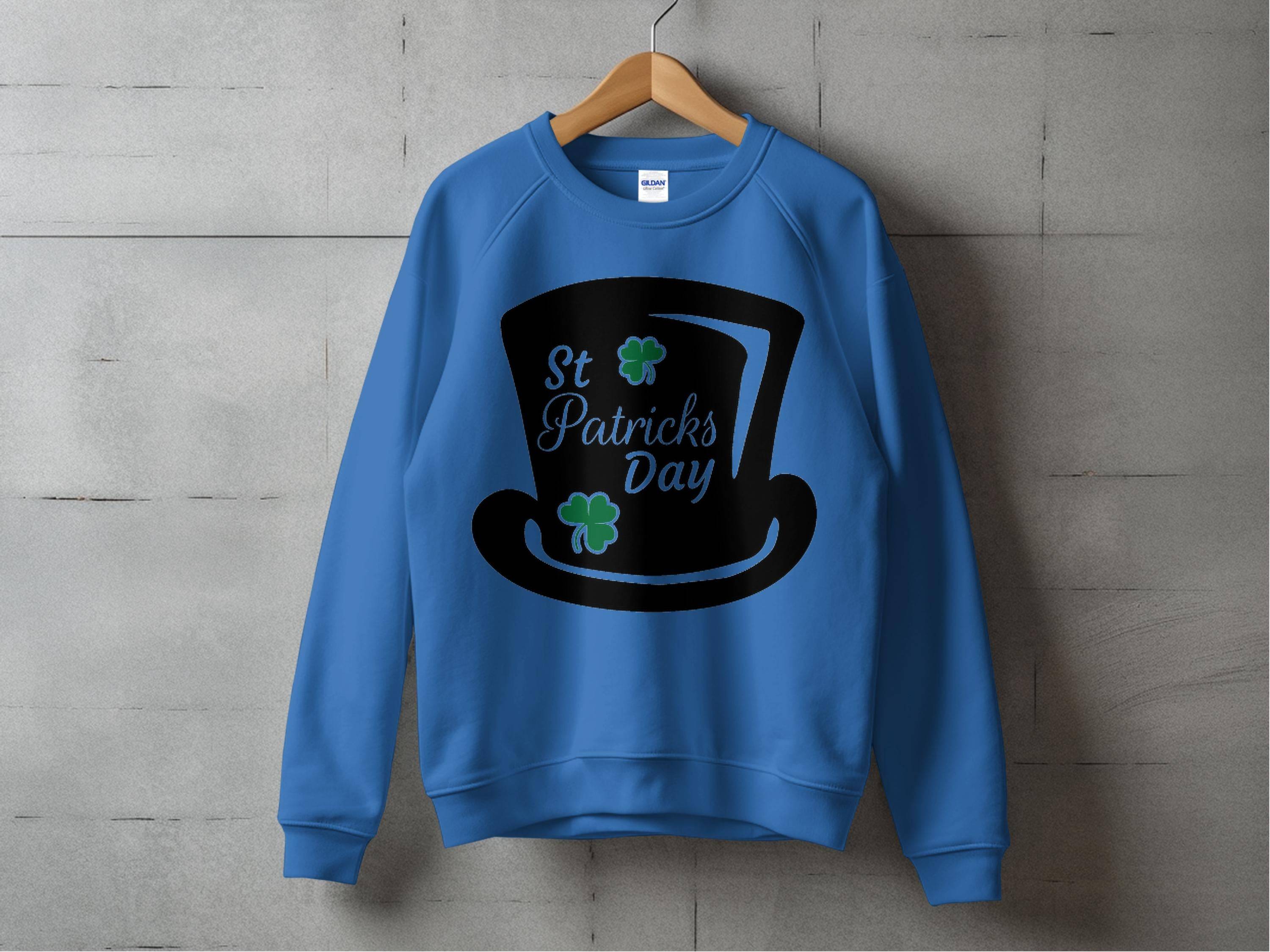 The Garment Graphics blue sweatshirt features a black top hat graphic with green clovers and St. Patricks Day text. Displayed on a wooden hanger against a concrete wall, it embodies festive charm for your celebrations.