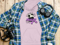 Flat lay image of a pink Soccer Mum hoodie featuring a soccer ball graphic, surrounded by a blue and green plaid shirt, black headphones, camera, and coffee cup on wood. The Garment Graphics hoodies offer a classic fit ideal for everyday style.