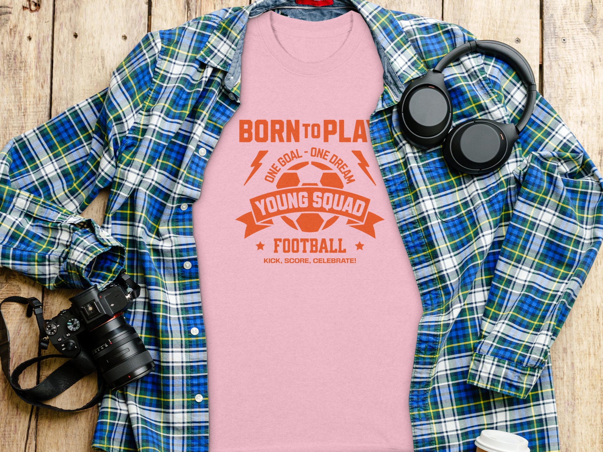 A Garment Graphics pink Born to Play, One Goal - One Dream football t-shirt rests on a wooden surface alongside a plaid shirt, headphones, and a camera.