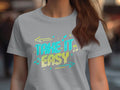 A person is wearing a Garment Graphics light gray T-shirt featuring the phrase Take It Easy in bright blue and yellow, accented by musical notes and arrows. The vibrant graphics enhance its trendy appeal against a blurred background.