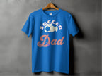 A blue graphic T-shirt from Garment Graphics hangs on a wooden hanger against a dark wall, showcasing the text SOCCER Dad with a soccer ball graphic above Dad.