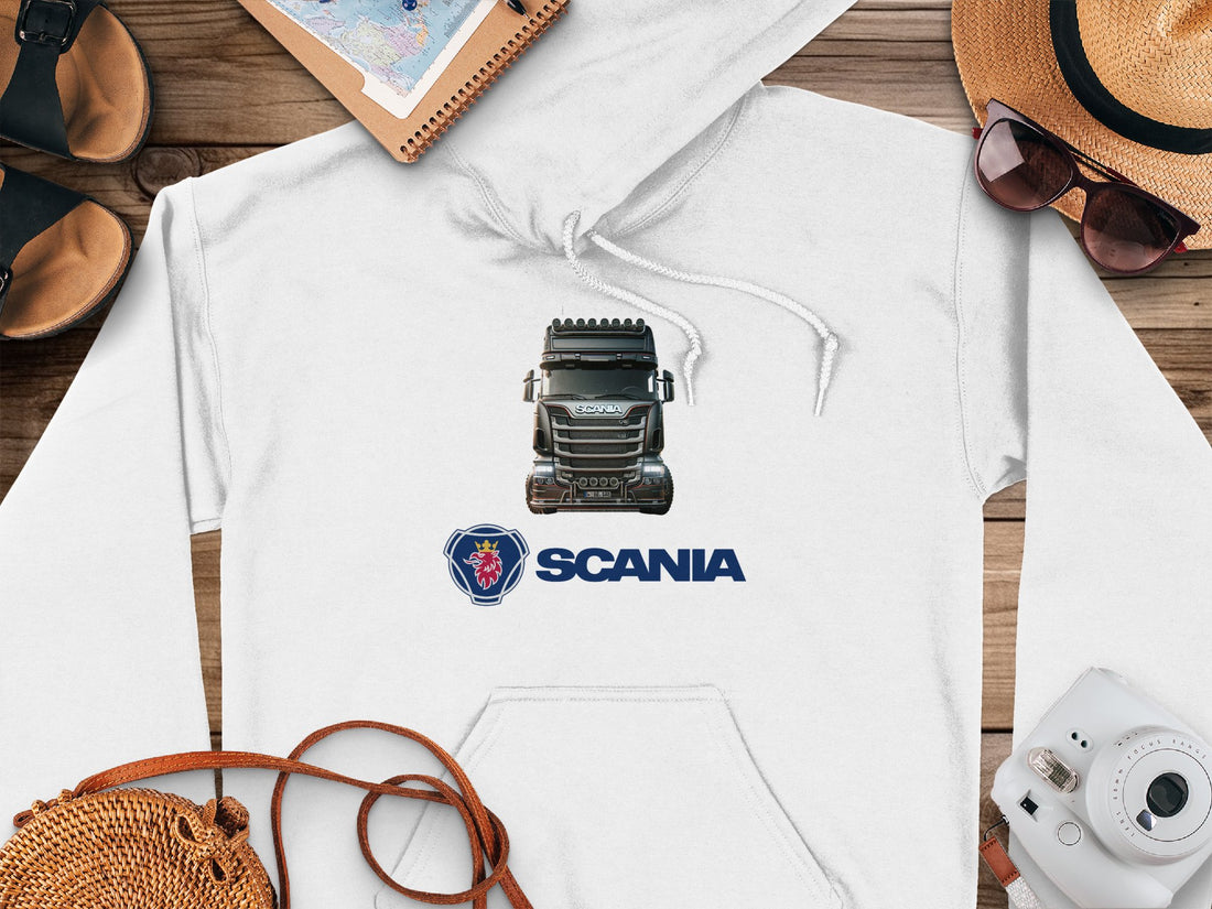 The Garment Graphics white hoodie features a graphic truck design with the Scania logo underneath. Ideal for truck enthusiasts, its displayed on a wooden surface alongside sandals, a notebook, sunglasses-topped straw hat, map, and camera.