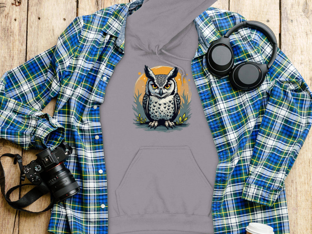 A flat lay features Garment Graphics gray owl artwork hoodie, made from medium-heavy fabric, elegantly arranged on a blue and green plaid shirt. Nearby are a camera, black headphones, and a white paper cup on a wooden surface.