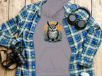 A flat lay features Garment Graphics gray owl artwork hoodie, made from medium-heavy fabric, elegantly arranged on a blue and green plaid shirt. Nearby are a camera, black headphones, and a white paper cup on a wooden surface.