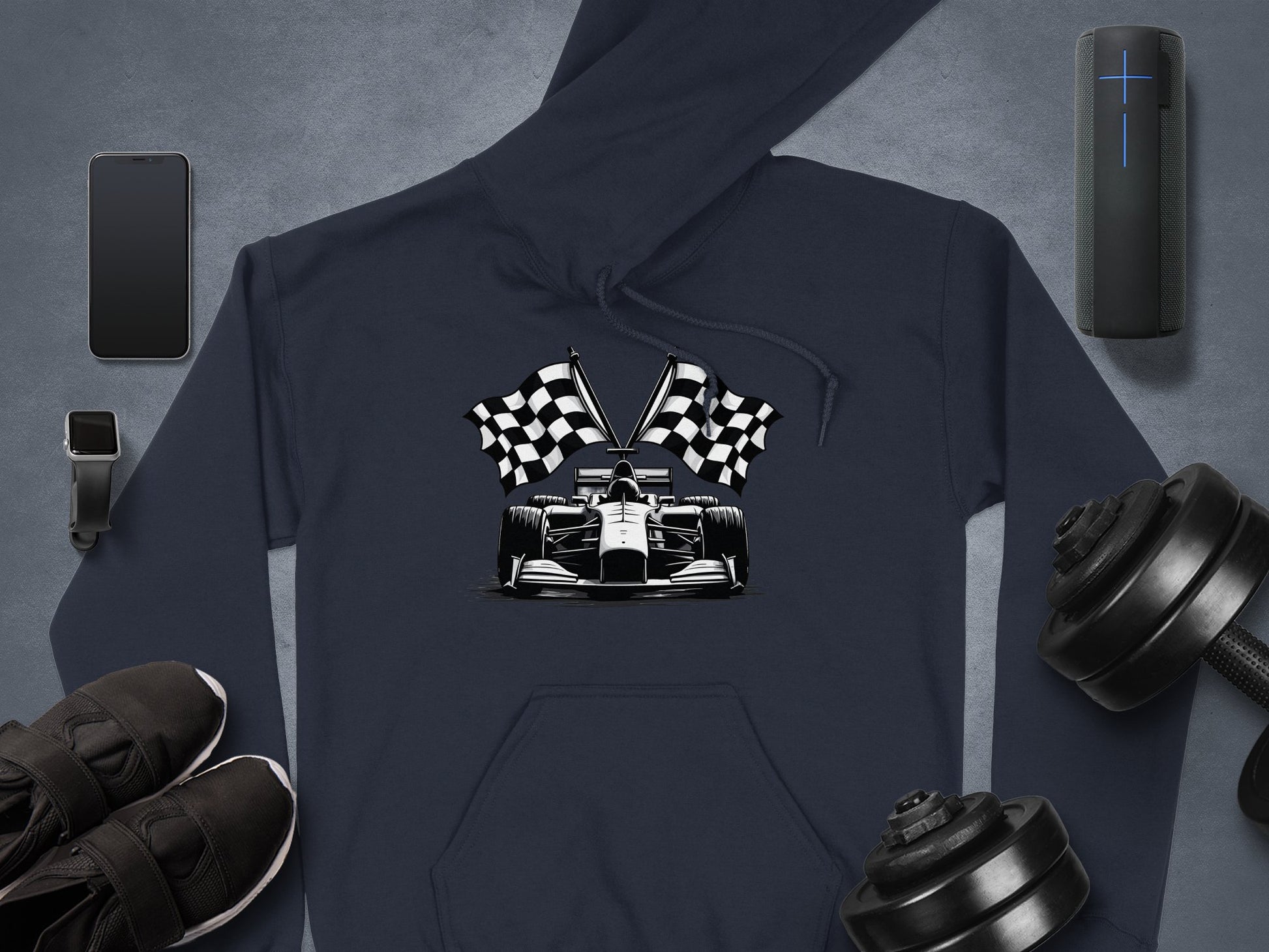 A Garment Graphics hoodie with a racing car and checkered flags design sits on a gray surface, surrounded by a smartphone, smartwatch, wireless speaker, sneakers, and adjustable dumbbells—ideal for motorsport fans.