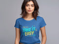 A person with medium-length brown hair is wearing a blue Garment Graphics Take It Easy T-Shirt adorned with vibrant graphics in a colorful, stylized font. The plain gray background complements the shirts stylish design.