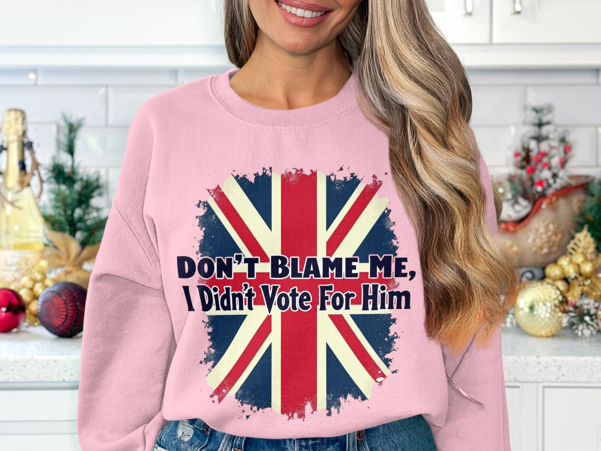 Amidst a festive kitchen, a woman wears a Garment Graphics pink sweatshirt with a distressed British flag and the phrase Dont Blame Me, I Didnt Vote For Him. Available in unisex sizes for versatile styling.