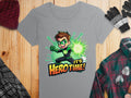 Garment Graphics offers a gray T-shirt with a green superhero design: a brown-haired cartoon character in a green suit, emitting a green energy burst. It features the text Its Hero Time! Displayed on a wooden surface with colorful plaid shirt, gloves, hat, and scarf.