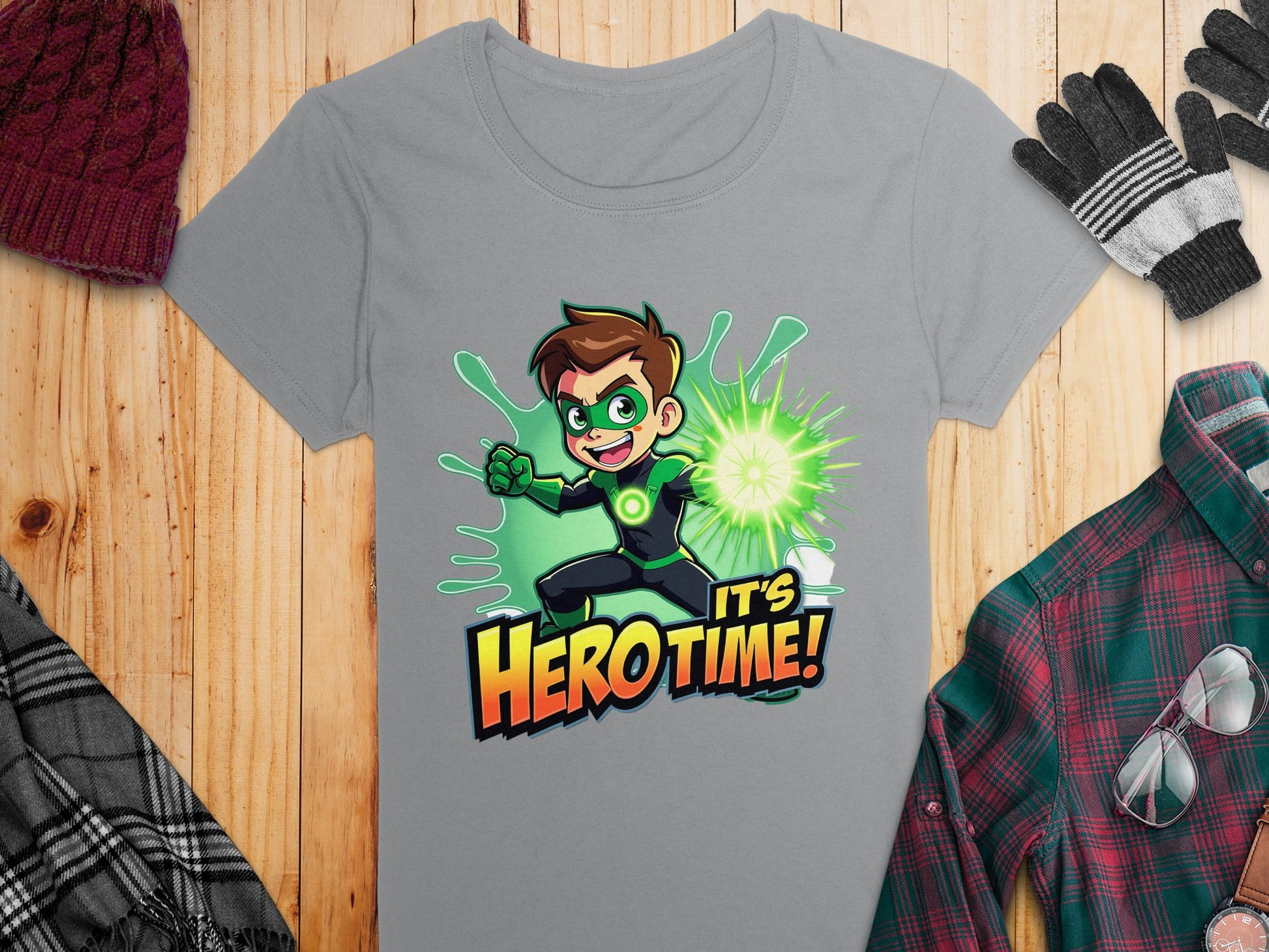 Garment Graphics offers a gray T-shirt with a green superhero design: a brown-haired cartoon character in a green suit, emitting a green energy burst. It features the text Its Hero Time! Displayed on a wooden surface with colorful plaid shirt, gloves, hat, and scarf.
