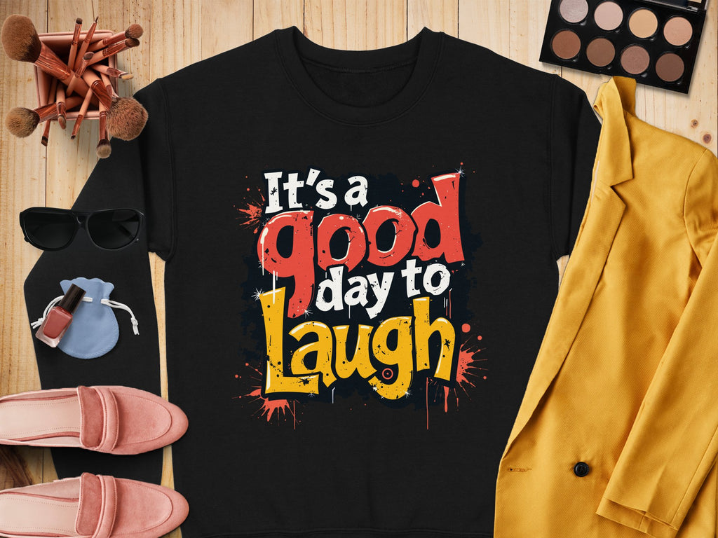 A black Garment Graphics sweatshirt with colorful Its a good day to laugh text, styled on wood with accessories: yellow jacket, pink shoes, makeup palette and brushes, sunglasses, and a blue pouch. Made from medium-heavy fabric for trendy unisex comfort.