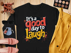 A black Garment Graphics sweatshirt with colorful Its a good day to laugh text, styled on wood with accessories: yellow jacket, pink shoes, makeup palette and brushes, sunglasses, and a blue pouch. Made from medium-heavy fabric for trendy unisex comfort.