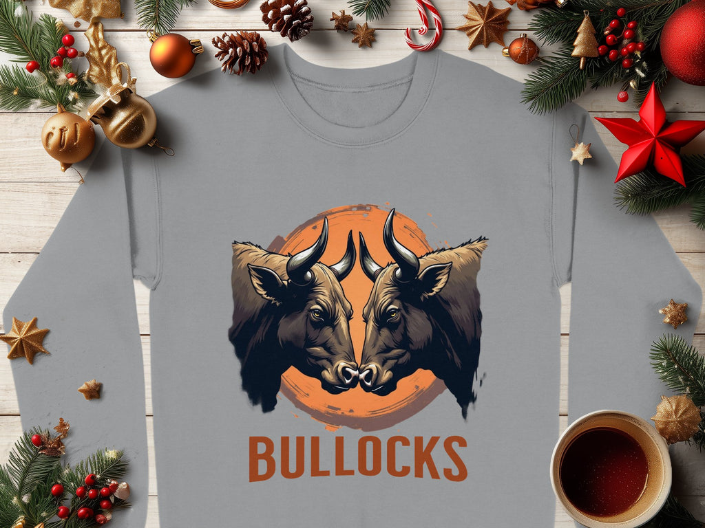 This Garment Graphics gray sweatshirt showcases a bold design with two bulls facing each other against an orange circle. BULLOCKS is printed below. Designed for a unisex fit, its framed by pine branches, ornaments, and a cup of coffee on wood.