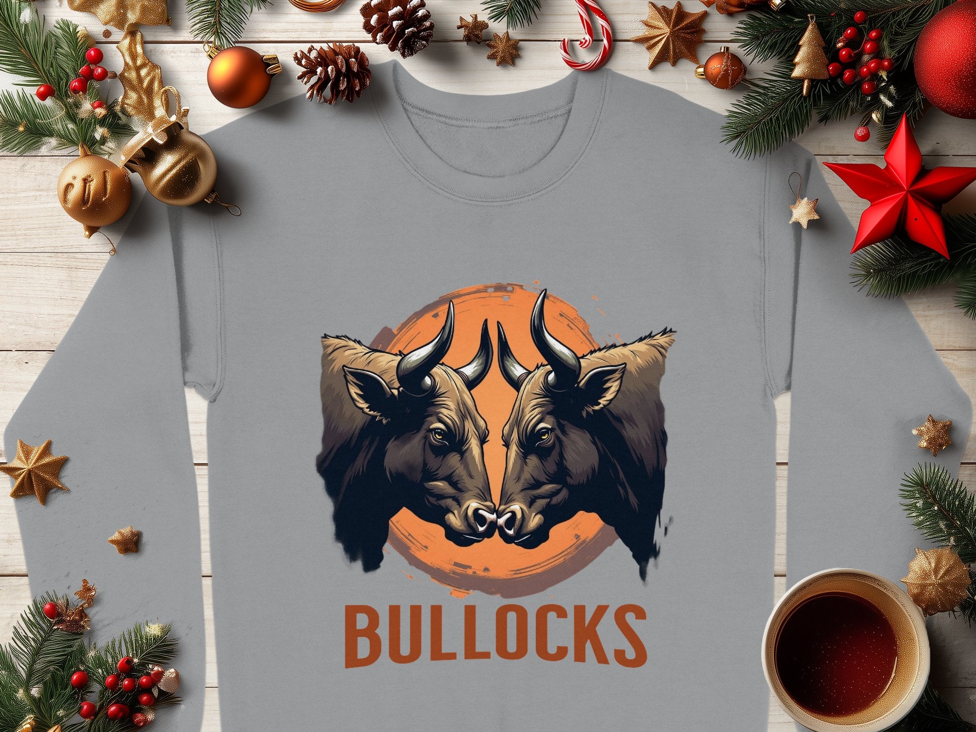 This Garment Graphics gray sweatshirt showcases a bold design with two bulls facing each other against an orange circle. BULLOCKS is printed below. Designed for a unisex fit, its framed by pine branches, ornaments, and a cup of coffee on wood.