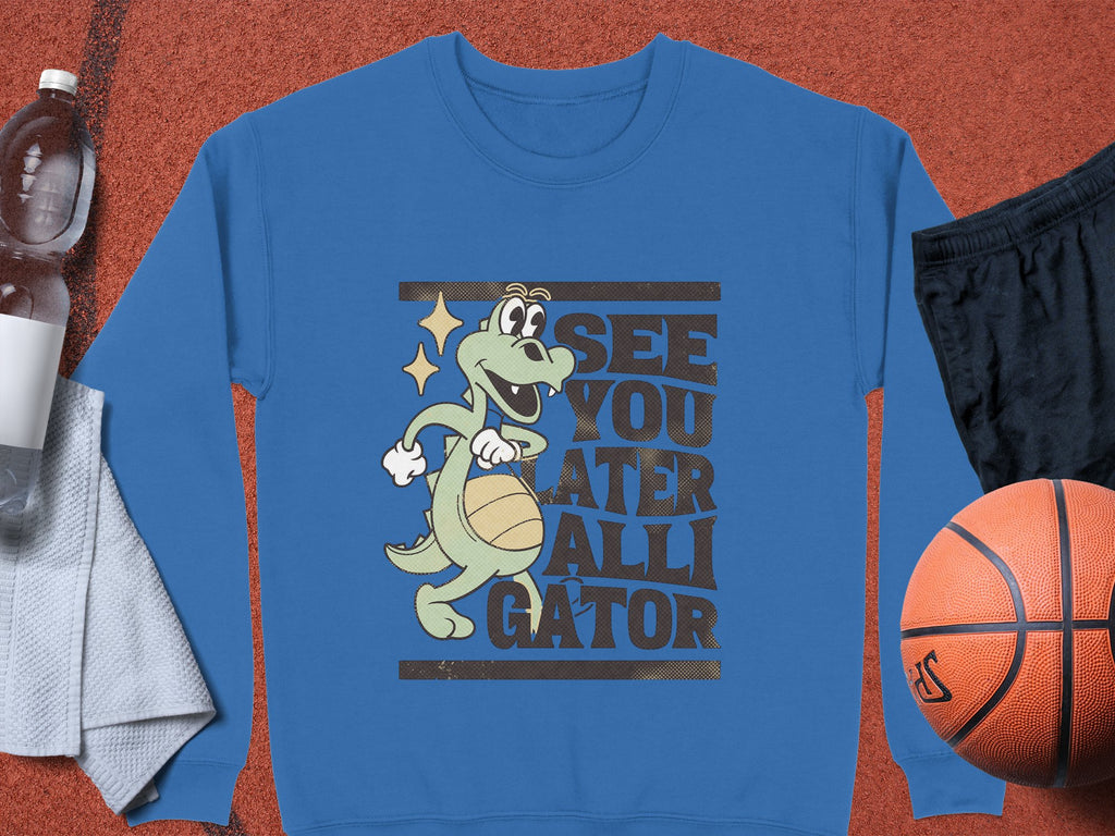 Garment Graphics offers a unisex sweatshirt featuring a cartoon alligator with a basketball and the phrase See You Later Alligator. Its pictured with a water bottle, light gray towel, black shorts, and basketball on an orange surface.