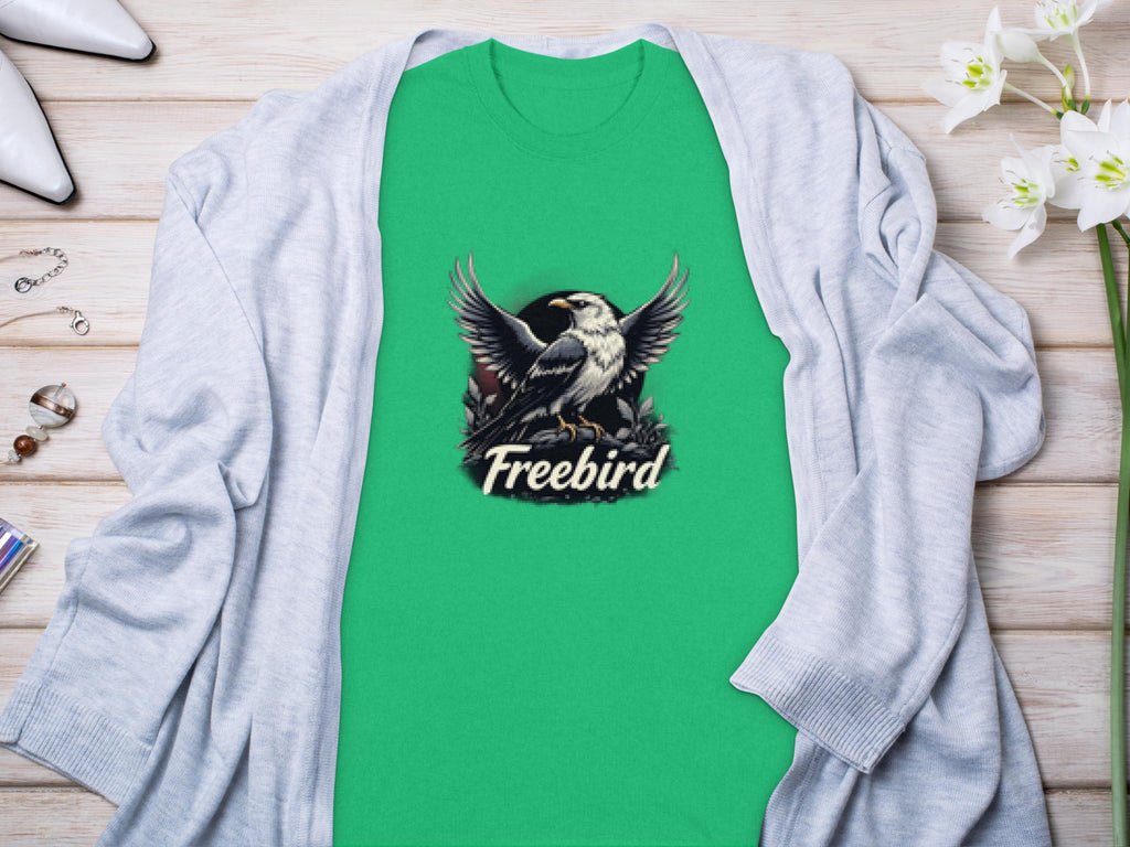 A Garment Graphics 100% cotton green t-shirt featuring a striking bird graphic and Freebird text lies elegantly on light wood, accessorized with a gray cardigan, white flowers, and chic items.