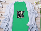 A Garment Graphics 100% cotton green t-shirt featuring a striking bird graphic and Freebird text lies elegantly on light wood, accessorized with a gray cardigan, white flowers, and chic items.