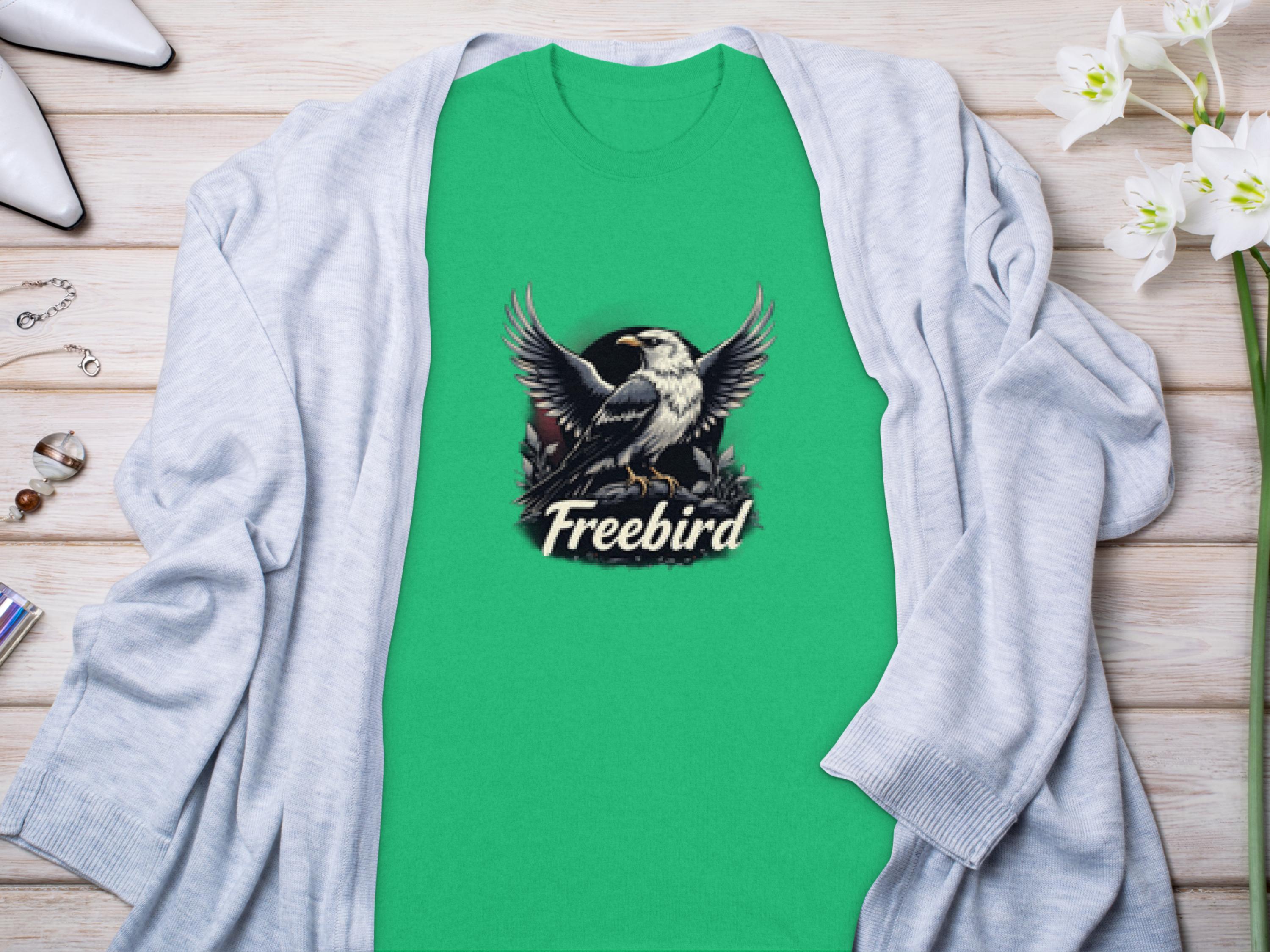 A Garment Graphics 100% cotton green t-shirt featuring a striking bird graphic and Freebird text lies elegantly on light wood, accessorized with a gray cardigan, white flowers, and chic items.