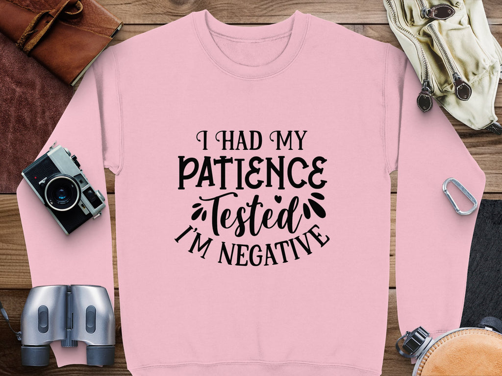 A pink Garment Graphics sweatshirt, laid flat on a wooden surface, displays the quirky slogan: I had my patience tested, Im negative. Ideal for casual outings, its surrounded by a camera, binoculars, gloves, and a leather pouch. Available in unisex sizes for added versatility.
