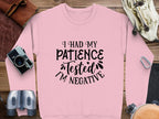 A pink Garment Graphics sweatshirt, laid flat on a wooden surface, displays the quirky slogan: I had my patience tested, Im negative. Ideal for casual outings, its surrounded by a camera, binoculars, gloves, and a leather pouch. Available in unisex sizes for added versatility.