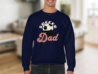 In the cozy living room, a person wears a Garment Graphics navy blue sweatshirt featuring the words Soccer Dad alongside a retro soccer ball design.