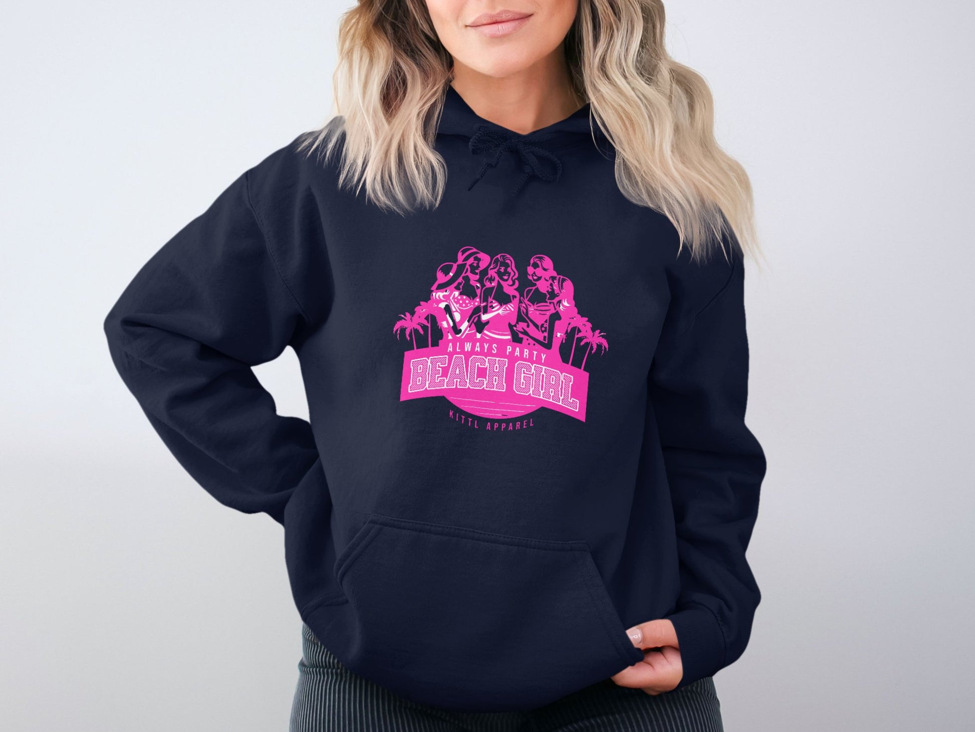 A woman is modeling the Garment Graphics Always Party Beach Girl hoodie, showcasing a casual style with a vibrant design of three silhouetted figures. Her attire features a navy blue backdrop and pink graphic that pops against a plain light gray background.